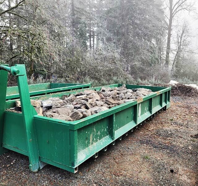 A green 10-yard dumpster, suitable for concrete and construction debris, parked on a gravel surface near a forested area with a partial view of a yellow excavator.
