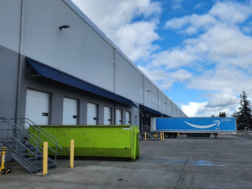 Large lime green roll-off dumpster rental parked at an Auburn, WA warehouse loading dock next to an Amazon Prime semi-truck.